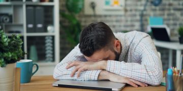 Tired office worker resting head on laptop at desk in a modern office.
