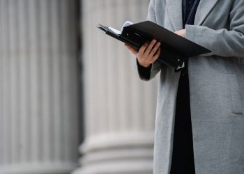 Crop unrecognizable businesswoman in gray coat standing on street with opened document folder and reading contract on autumn day