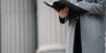 Crop unrecognizable businesswoman in gray coat standing on street with opened document folder and reading contract on autumn day