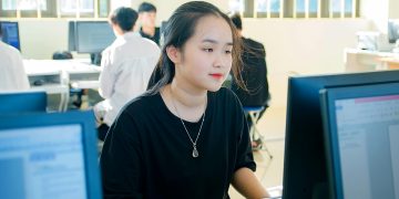 A young Asian woman concentrating on a computer screen in a university classroom.