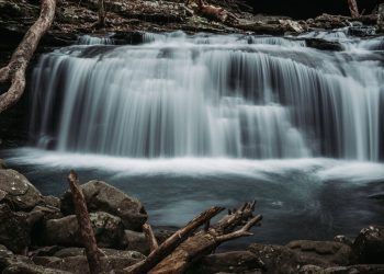A serene view of a cascading waterfall in a forest, surrounded by rocks and logs, capturing the essence of nature.