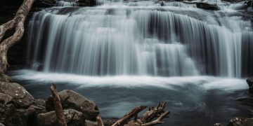 A serene view of a cascading waterfall in a forest, surrounded by rocks and logs, capturing the essence of nature.