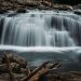 A serene view of a cascading waterfall in a forest, surrounded by rocks and logs, capturing the essence of nature.