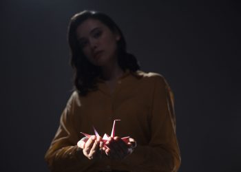 A moody portrait of a woman holding an origami paper crane in low light.