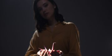 A moody portrait of a woman holding an origami paper crane in low light.
