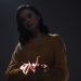 A moody portrait of a woman holding an origami paper crane in low light.