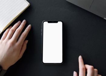 A minimalist workspace featuring a smartphone mockup, hands, and a notebook on a black surface.