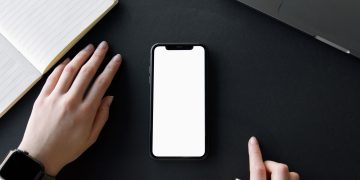 A minimalist workspace featuring a smartphone mockup, hands, and a notebook on a black surface.