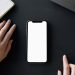 A minimalist workspace featuring a smartphone mockup, hands, and a notebook on a black surface.