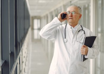 Serious mature male doctor in medical uniform standing with stethoscope and notebook while talking on smartphone in clinic corridor