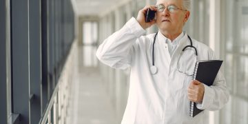 Serious mature male doctor in medical uniform standing with stethoscope and notebook while talking on smartphone in clinic corridor