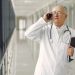 Serious mature male doctor in medical uniform standing with stethoscope and notebook while talking on smartphone in clinic corridor
