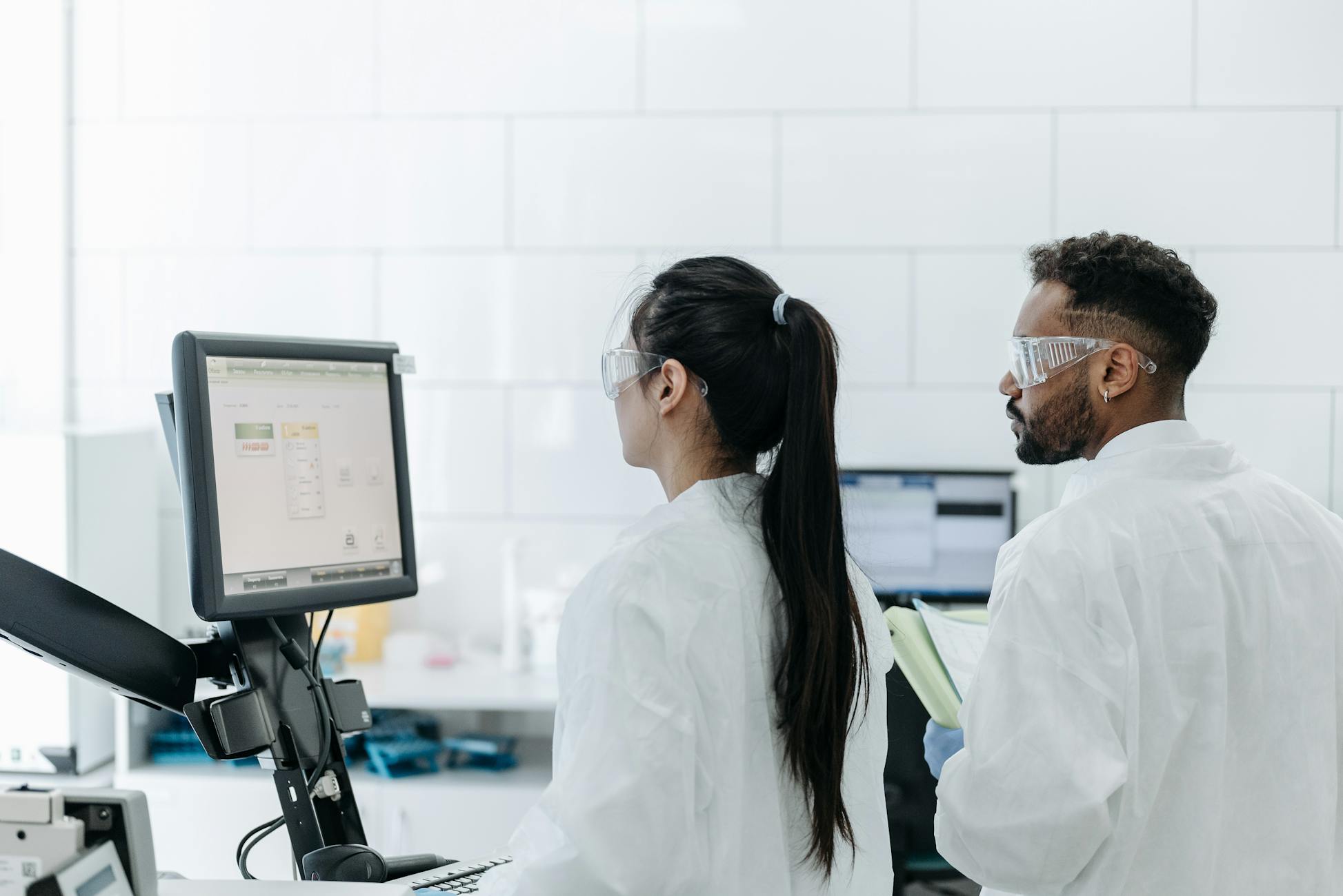Two scientists wearing lab coats and goggles analyze data on a computer in a modern laboratory.
