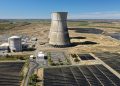 Aerial view of a nuclear cooling tower surrounded by solar panel fields in a rural landscape.