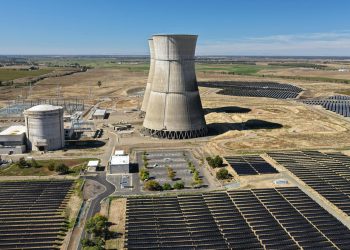 Aerial view of a nuclear cooling tower surrounded by solar panel fields in a rural landscape.