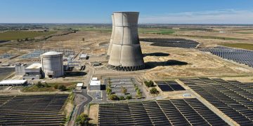 Aerial view of a nuclear cooling tower surrounded by solar panel fields in a rural landscape.
