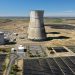 Aerial view of a nuclear cooling tower surrounded by solar panel fields in a rural landscape.