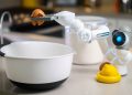 A robotic helper cracks an egg into a bowl in a contemporary kitchen setting, showcasing automation in cooking.