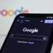Smartphone screen showing Google search in dark mode with the Google logo in the background.