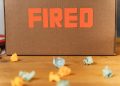 Close-up of a cardboard box labeled FIRED on a wooden table with crumpled papers.