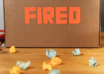 Close-up of a cardboard box labeled FIRED on a wooden table with crumpled papers.