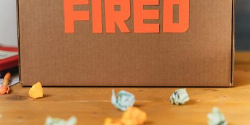Close-up of a cardboard box labeled FIRED on a wooden table with crumpled papers.