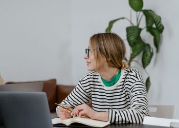 A woman in glasses sits at a desk, writing in a notebook with a laptop nearby, indoors.
