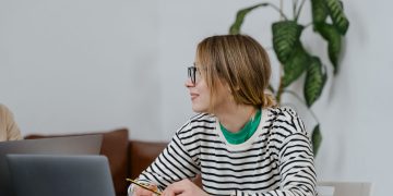 A woman in glasses sits at a desk, writing in a notebook with a laptop nearby, indoors.