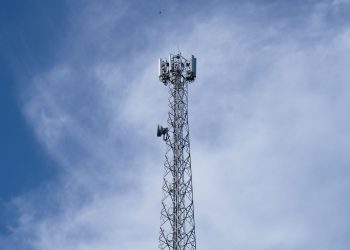 A tall metal communications tower stands against a backdrop of a clear blue sky with scattered clouds.