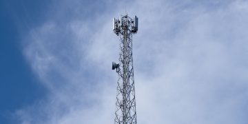 A tall metal communications tower stands against a backdrop of a clear blue sky with scattered clouds.