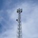 A tall metal communications tower stands against a backdrop of a clear blue sky with scattered clouds.