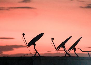 A row of satellite dishes silhouetted against a vibrant orange sunset sky on a rooftop.