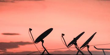 A row of satellite dishes silhouetted against a vibrant orange sunset sky on a rooftop.