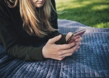 A young woman using her smartphone while lying on a blanket outdoors.