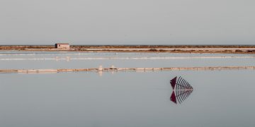 A tranquil scene of salt flats with still water reflecting structures under a clear sky.