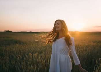A woman in a white dress smiles joyfully amidst a sunset-lit wheat field.