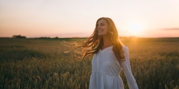 A woman in a white dress smiles joyfully amidst a sunset-lit wheat field.