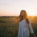 A woman in a white dress smiles joyfully amidst a sunset-lit wheat field.