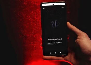 A hand holds a smartphone displaying Grok 3 announcement against a red background.
