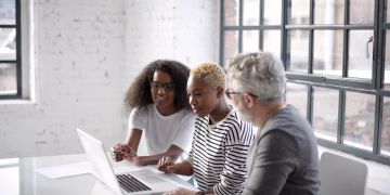 Side view of black women and old gray haired man sitting at table and surfing laptop while working on creative project