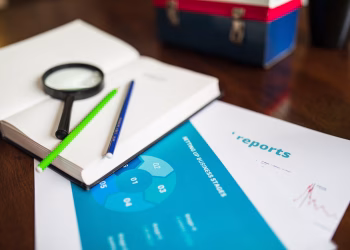 Open book with magnifying glass, pencils, and a business report on a wooden desk.