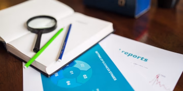 Open book with magnifying glass, pencils, and a business report on a wooden desk.