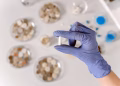 Close-up of a gloved hand holding a white capsule in a lab environment.
