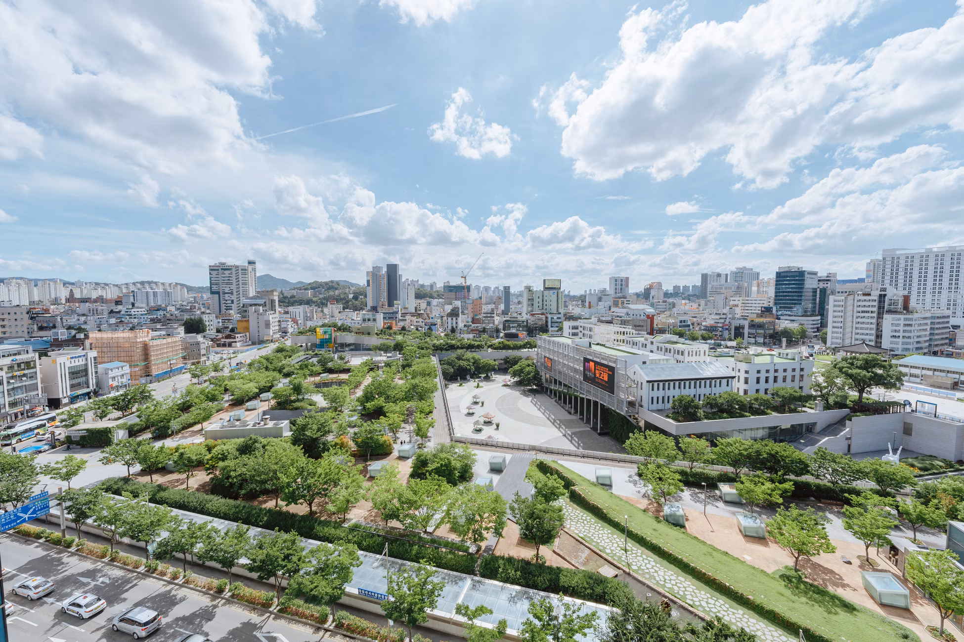 A vibrant cityscape featuring a lush urban park surrounded by modern buildings under a partly cloudy sky.