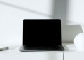 A sleek black laptop on a white desk in a minimalist setup with natural lighting.