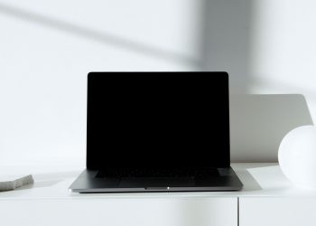 A sleek black laptop on a white desk in a minimalist setup with natural lighting.