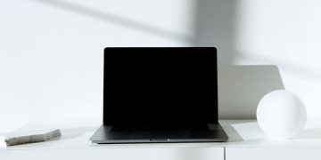 A sleek black laptop on a white desk in a minimalist setup with natural lighting.