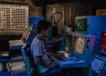 A man working at control panels in a dimly lit industrial room with various monitors.