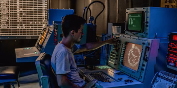A man working at control panels in a dimly lit industrial room with various monitors.