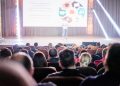 Audience attentively watching a presentation in a conference setting, with stage lights and speaker visible.